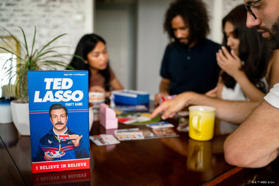 De doos van het partyspel Ted Lasso en mensen op de achtergrond die het spel spelen